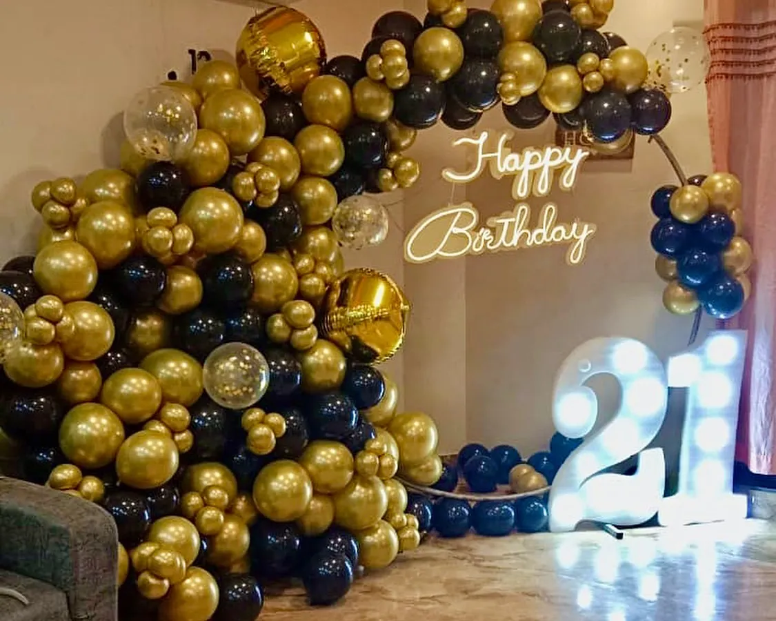 Celebrating a 21st birthday in style. This setup features a dramatic black and gold organic balloon arch, a custom 'Happy Birthday' neon sign, and large, illuminated marquee numbers to mark the special occasion in any home.
