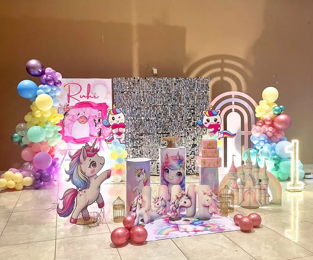 A magical unicorn-themed first birthday party. This setup features a sparkling sequin backdrop, pastel rainbow balloon garlands, and custom props including unicorn cutouts and themed plinths for the cake display. The large, light-up number '1' adds a personalized, photo-ready touch.