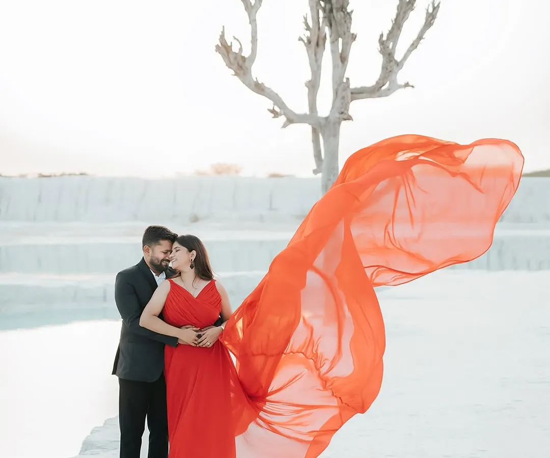 A bold and dramatic shot from a pre-wedding session at a mineral quarry. The vibrant red of the flowing gown against the stark white landscape creates a powerful, high-fashion image that tells a story of passionate love.