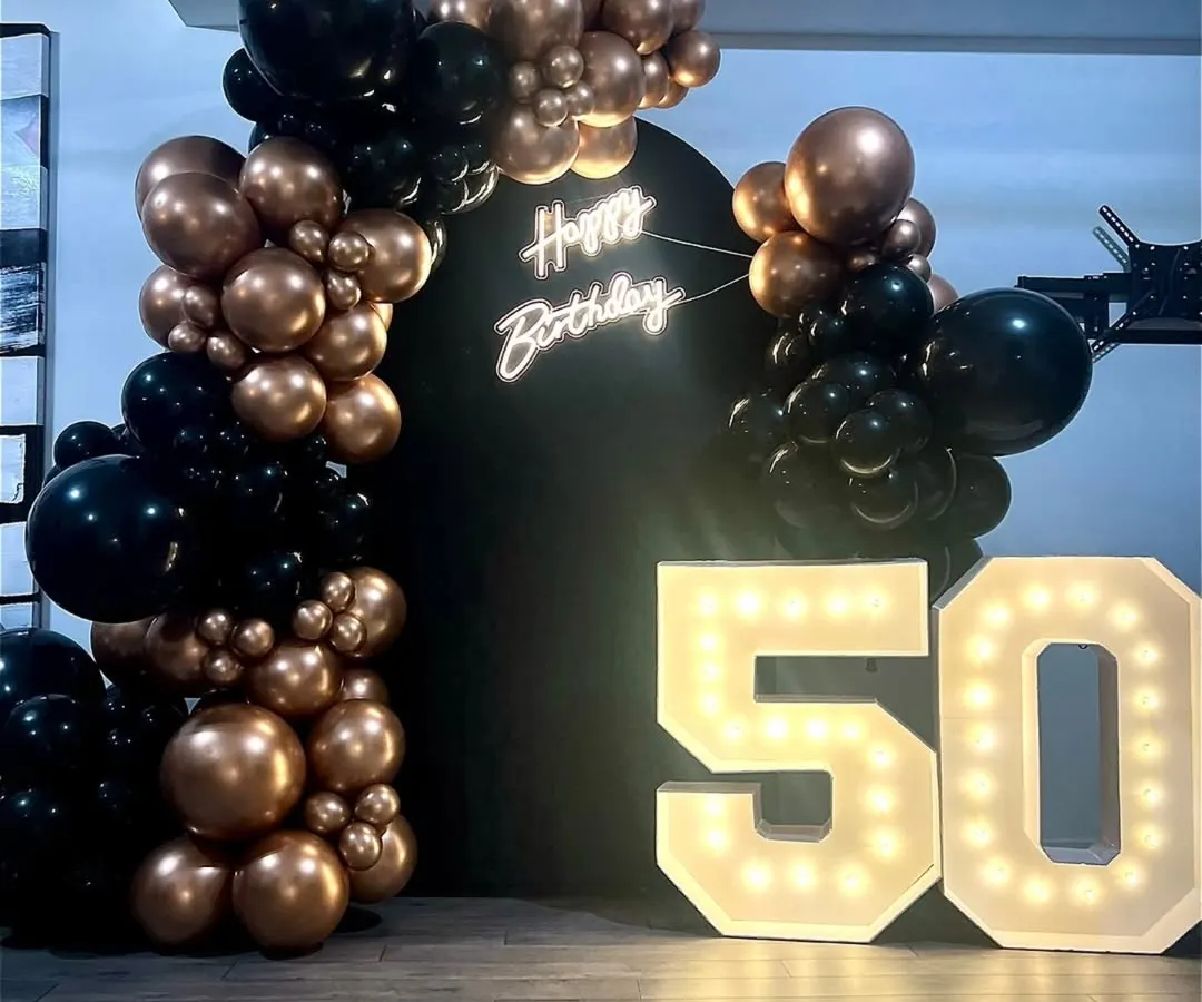 A chic and modern 50th birthday celebration. The setup combines a black backdrop with a rose gold balloon arch, a "Happy Birthday" neon sign, and large marquee numbers for a sophisticated and glamorous party vibe.