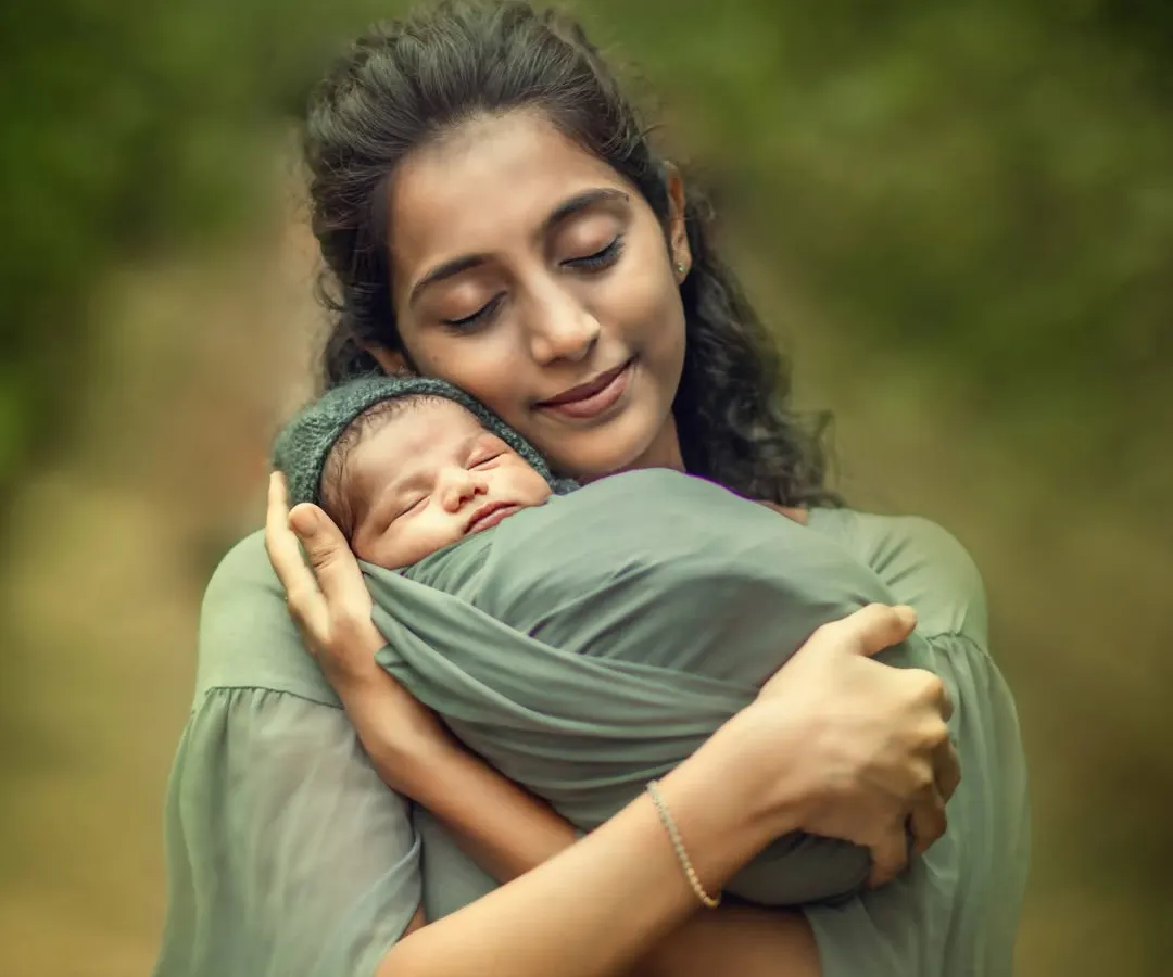 A new mother, with her eyes gently closed, cradles her sleeping, swaddled newborn against a soft, green forest backdrop. This outdoor newborn portrait captures the pure serenity and overwhelming love of early motherhood.