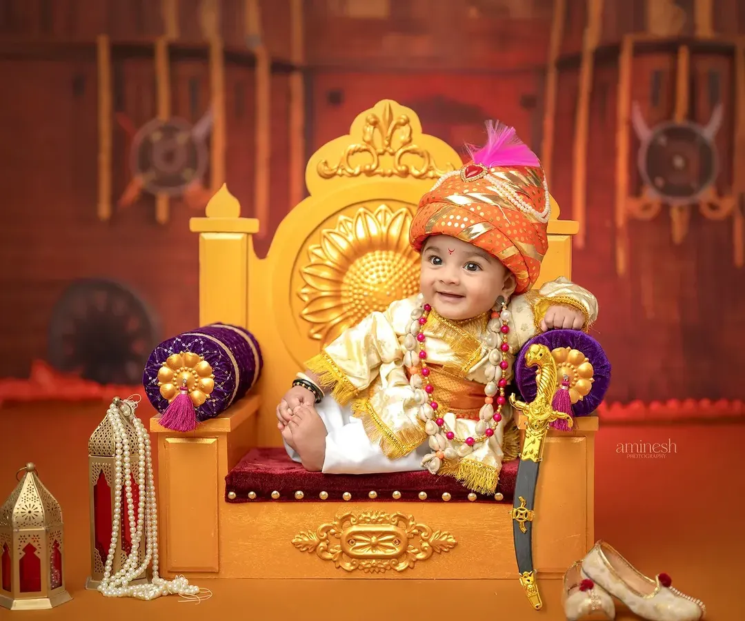 Capturing the spirit of a young king, this Bal Shivba themed portrait features a six-month-old baby in royal attire on a miniature throne. The setup includes a toy sword and traditional shoes, set against a backdrop of a Maratha fort.