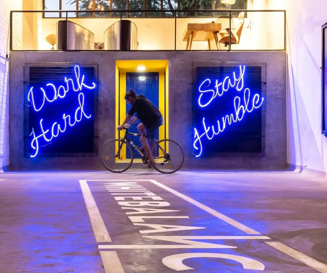 We transformed an old warehouse into an energetic office space, using neon signs like 'Work Hard Stay Humble' to motivate and inspire. The open-plan design includes unique features like an indoor cycling track, blending industrial grit with a playful, modern spirit to create a workspace that's anything but ordinary.