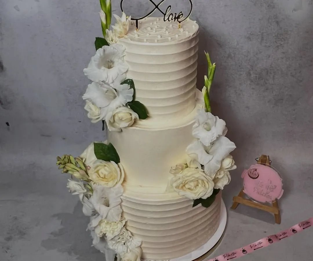 A timeless two-tier wedding cake with a classic ridged buttercream finish. It's adorned with fresh white gladiolus and roses, and topped with an infinity 'Love' sign, perfect for an elegant and traditional celebration.