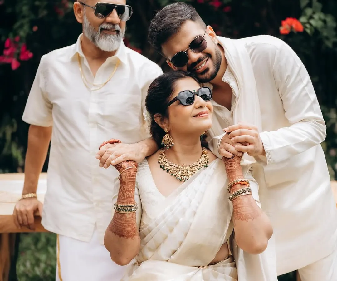 A modern family portrait, captured outdoors with natural light. The son embraces his mother, who is seated, while the father stands beside them, all sharing a moment of relaxed connection in coordinated white outfits.