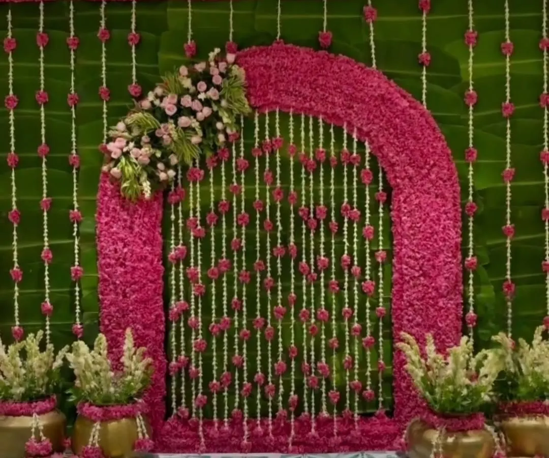 For this Ganpati celebration, I designed a vibrant floral arch using pink blossoms against a lush green backdrop of banana leaves. The hanging strings of jasmine and roses add a delicate, traditional touch.