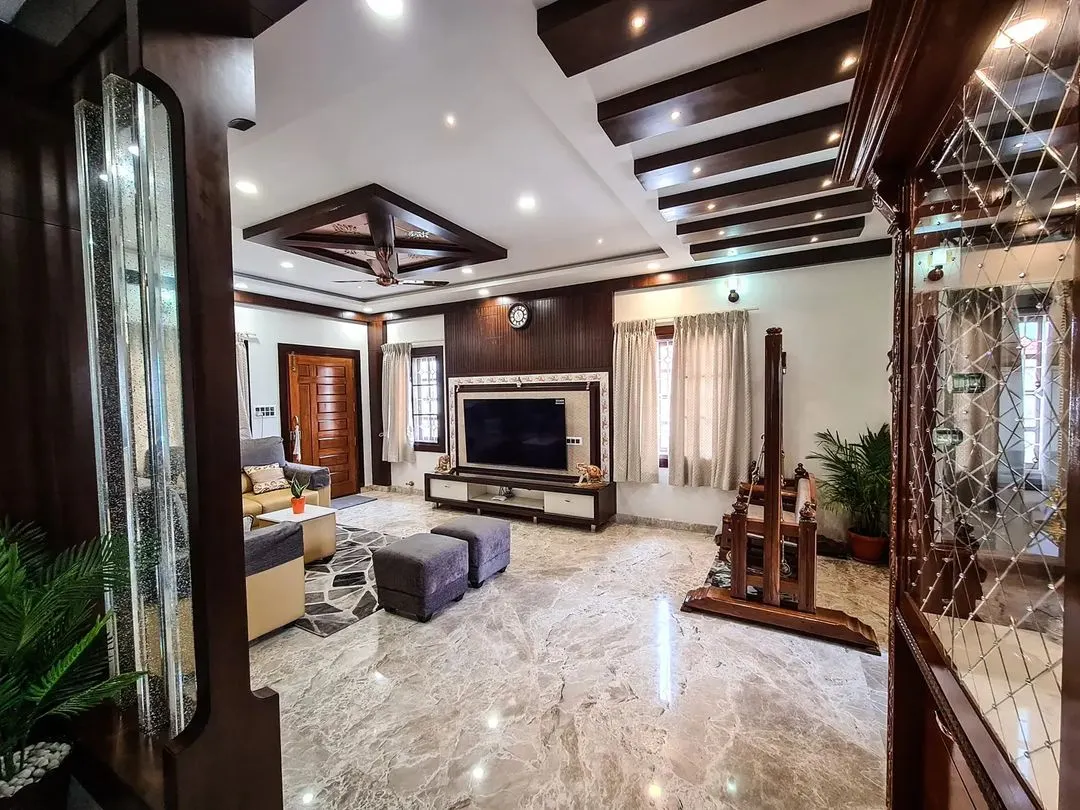I designed this expansive living room to feel both grand and inviting, featuring Italian marble flooring and a complex coffered ceiling with both wood beams and recessed lighting. The space is anchored by a large, modern TV unit and includes a traditional wooden jhoola (swing), blending contemporary luxury with classic Indian elements.