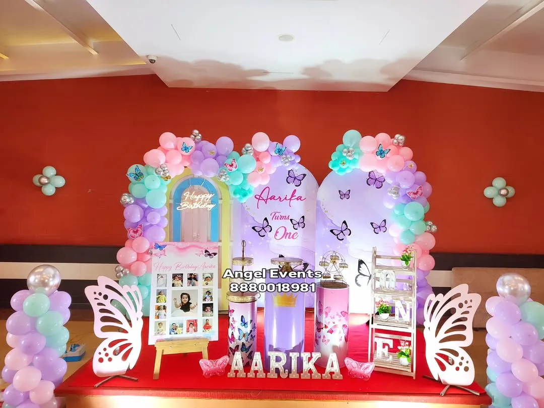 An enchanting butterfly theme in pastel shades of purple, pink, and mint green. The decoration features two arched backdrops, a full balloon garland, custom name lettering, and whimsical butterfly wing props for a magical party atmosphere.
