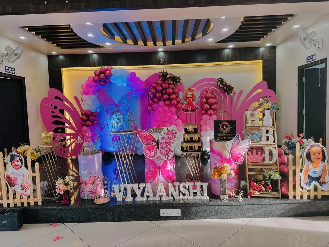 An elaborate stage setup for a butterfly-themed birthday party. This grand design features large pink butterfly wing cutouts, balloon garlands, custom-lit name signs, and themed pedestals for the cake, transforming the venue into a fairytale setting.