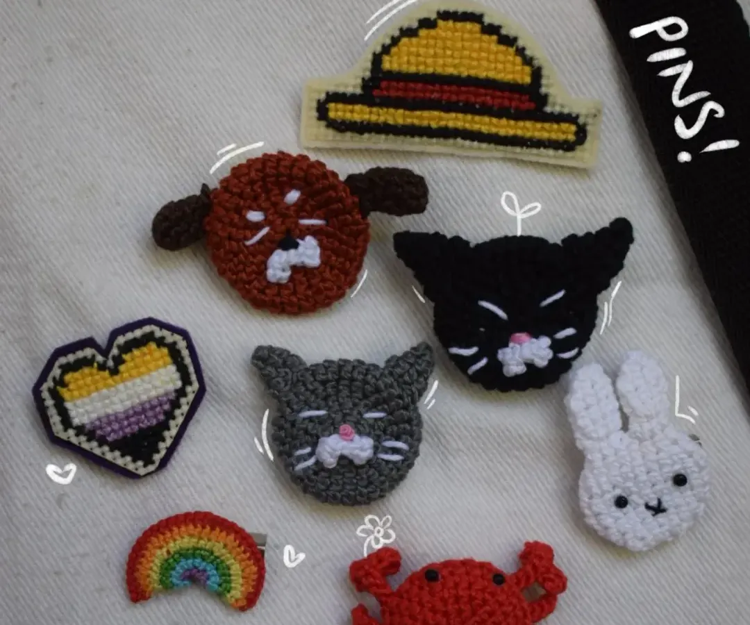 A collection of my cool handmade pins laid out on a tote bag. You can see the variety I offer, from a non-binary pride flag heart and an ace pride flag heart to cute animals like a black cat and a little crab. There's even a straw hat pin for all you anime fans.