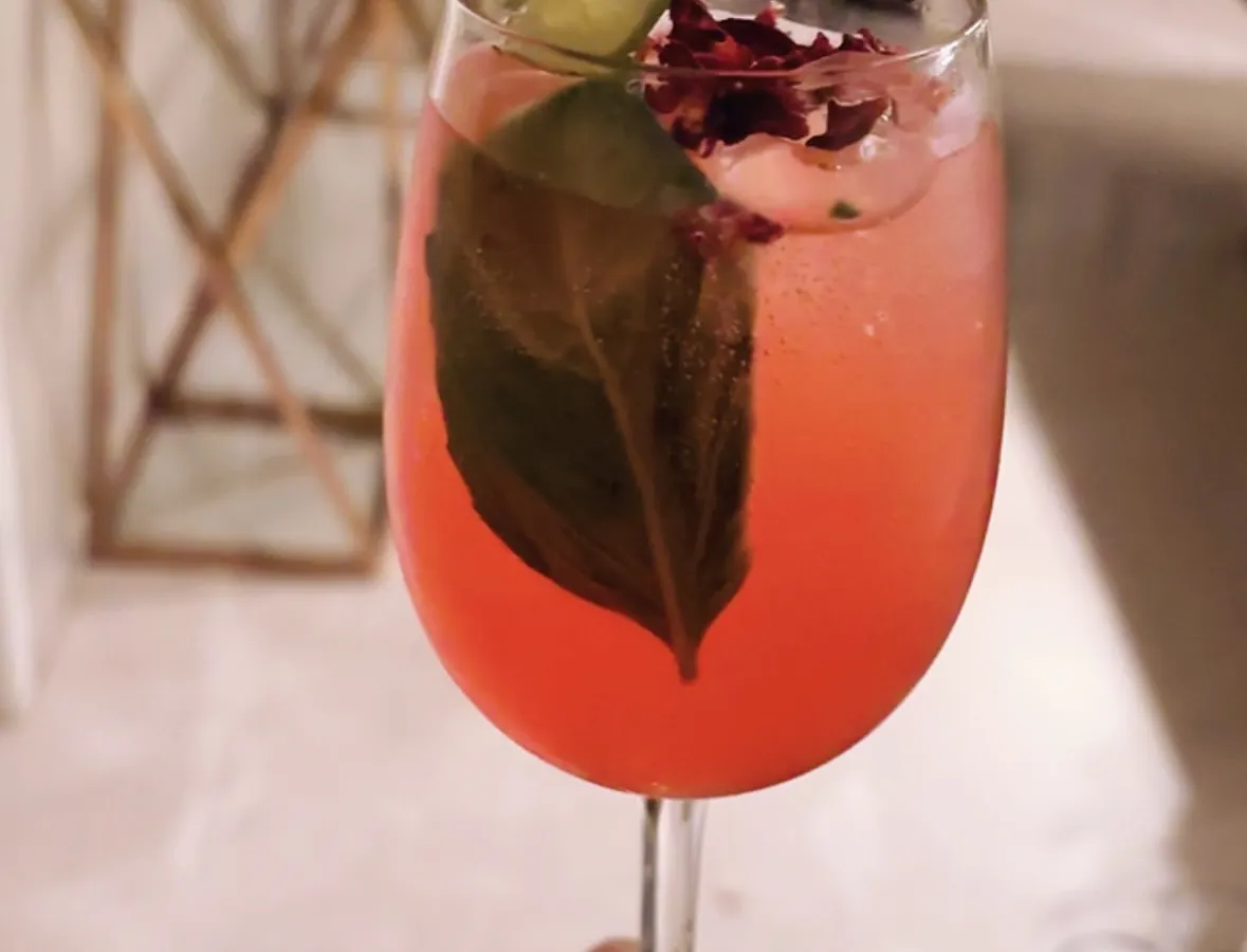 A beautifully crafted signature gin cocktail, showcasing our attention to detail. We use fresh ingredients like basil and rose petals, with a lime wheel garnish, to create drinks that are as visually appealing as they are delicious.