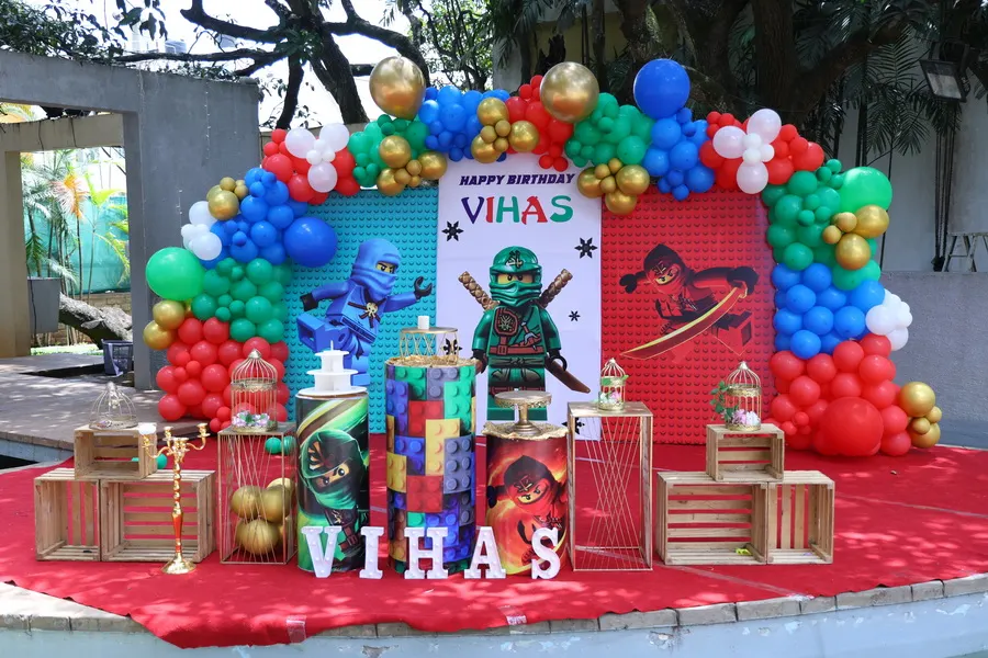 A complete birthday party setup for a Ninjago theme, featuring a custom backdrop, character displays, themed plinths for cake and desserts, and a vibrant balloon arch.