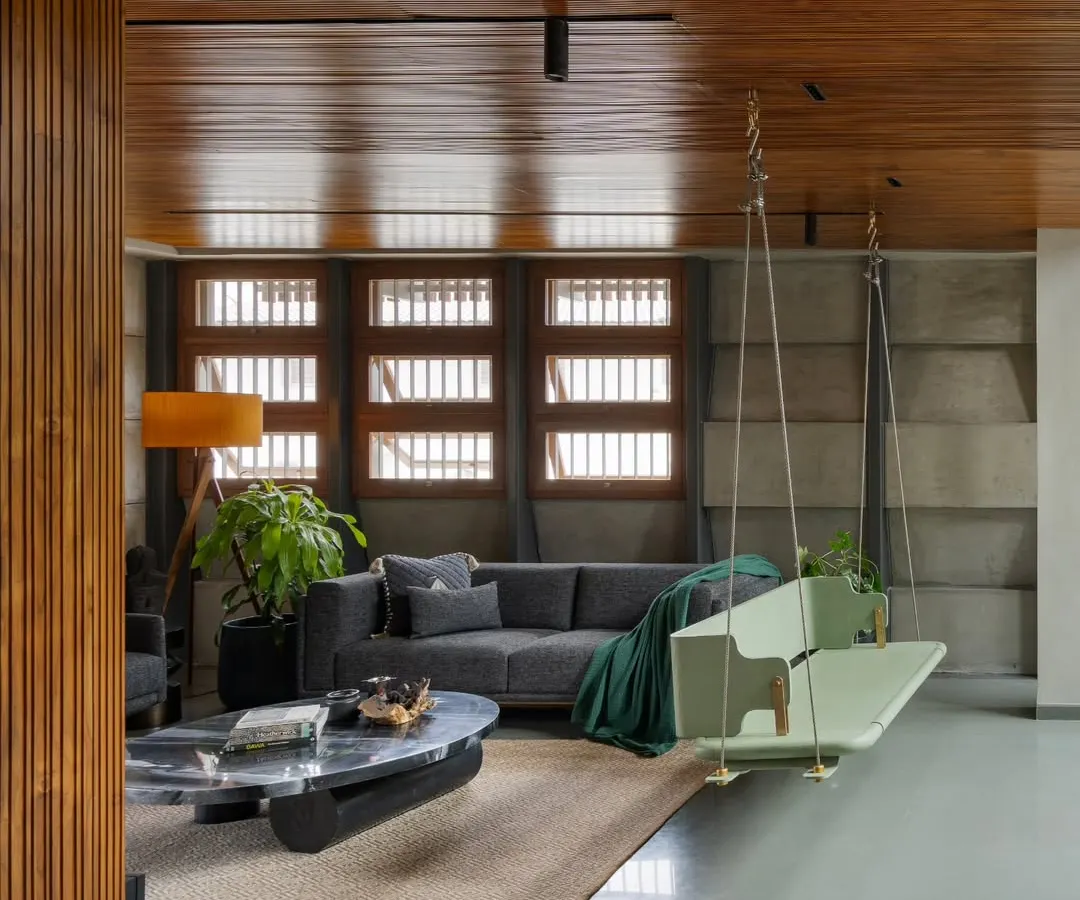 The living area of the 'Heirlooms in Concrete' home, where a contemporary swing adds a playful element. We combined a warm wood ceiling, textured concrete walls, and polished grey flooring to create a space that feels both expansive and intimate.