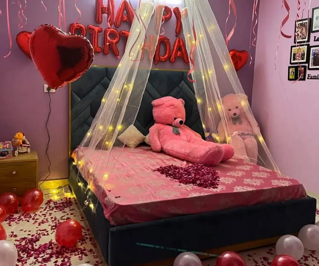 A cozy and romantic birthday room surprise. The setup features a bed with a light-up canopy, "HAPPY BIRTHDAY" foil balloons, a large teddy bear, and the floor covered in red and white balloons with rose petals.