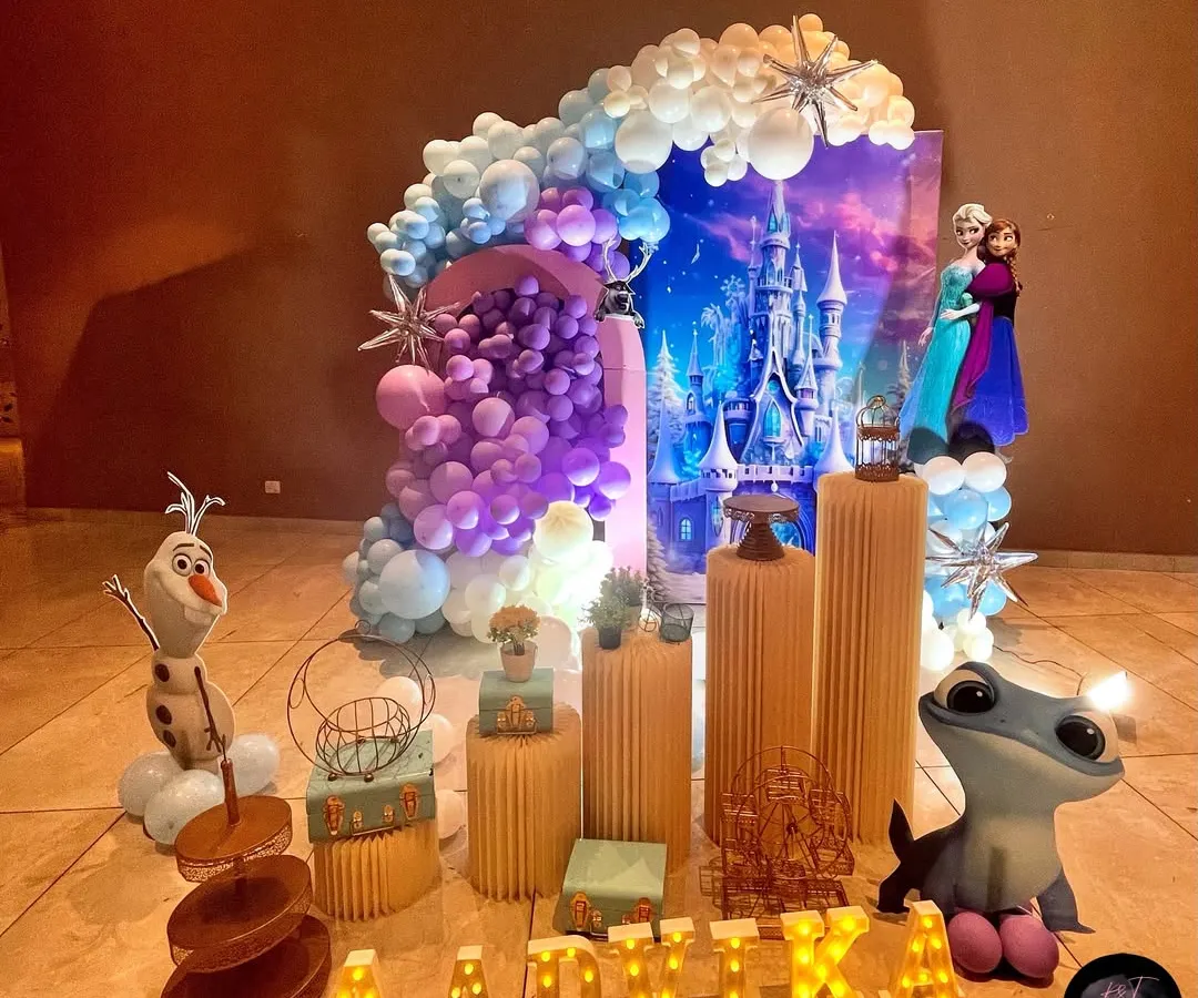 A magical Disney 'Frozen' party setup for Aadvika. This wide shot shows the stunning Arendelle castle backdrop, an elaborate balloon arch in shades of purple, blue, and white, and character cutouts that bring the winter wonderland to life.