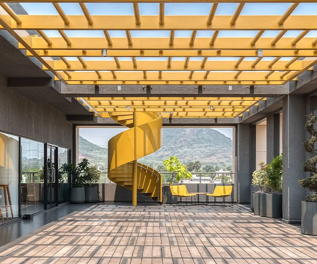 A bold, yellow spiral staircase serves as a sculptural centerpiece on the terrace of the Laurus Labs regional office in Vizag. This open-air space was designed to maximize natural light and offer stunning views of the surrounding hills, creating an inspiring breakout area that encourages movement and connection with the outdoors.