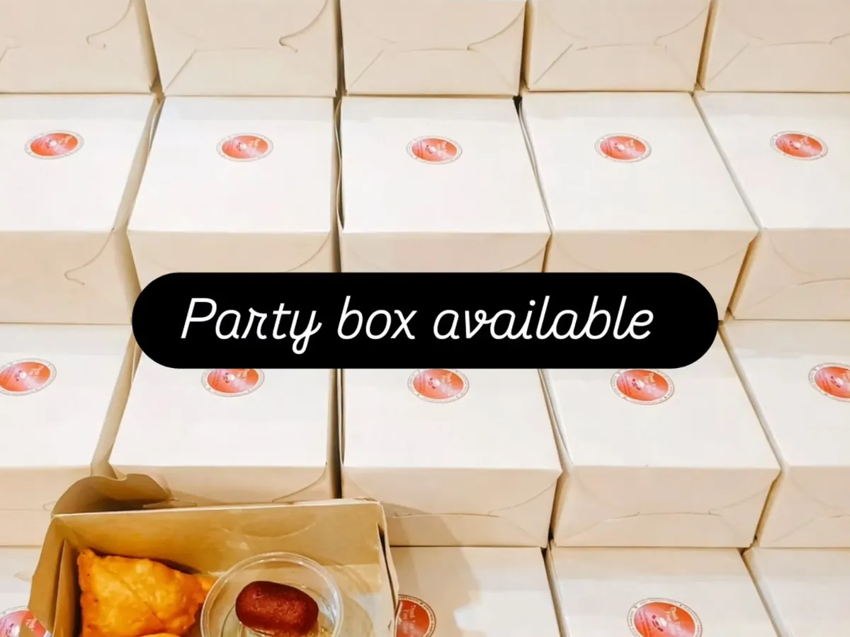 Samosa, a sweet and chutneys sit in an open snack box beside the closed party boxes we have packed.