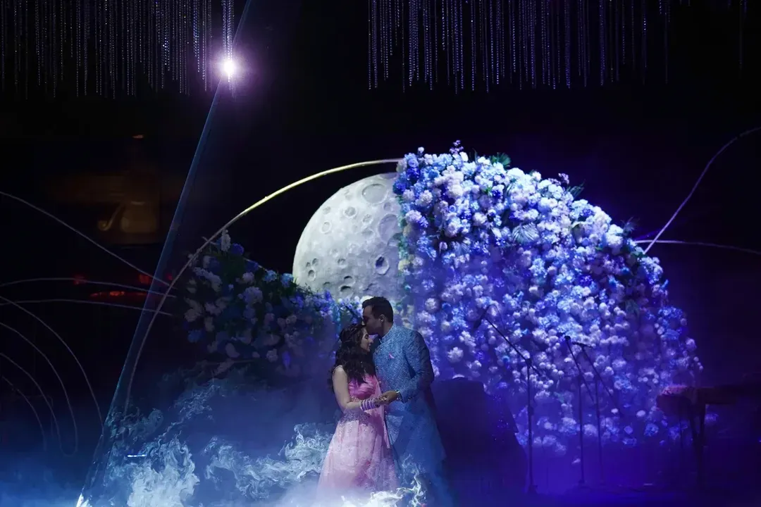 A magical moment as the couple shares a kiss under a stunning floral moon installation, part of our 'Starry Night' engagement theme.