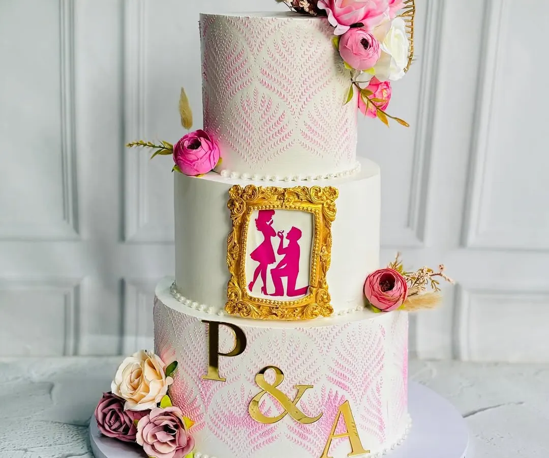 I love telling a love story through cake. This stunning three-tier engagement cake features a romantic proposal silhouette inside a golden frame, delicate stencil work, and beautiful florals, capturing the magic of a "yes" moment.