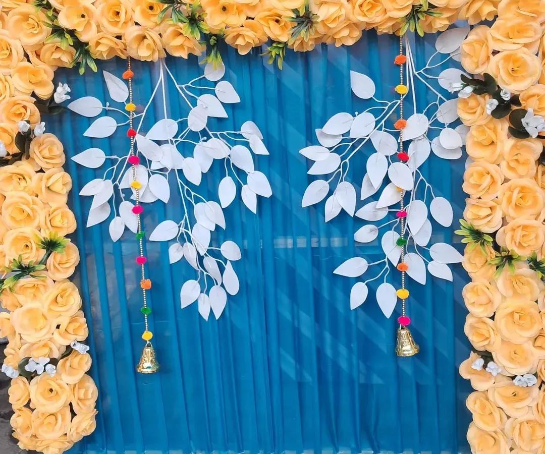 For a festive pooja backdrop, I combined a bright yellow artificial rose frame with a rich blue fabric curtain. The design is accented with decorative white leaf hangings and small bells for a unique and modern look.