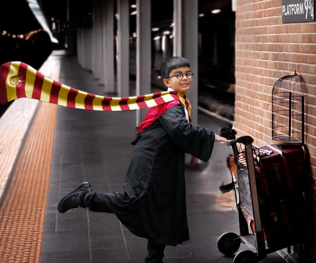 A magical Harry Potter themed photoshoot featuring a young boy in a Gryffindor robe pushing a luggage trolley towards a Platform 9¾ sign on a brick wall.