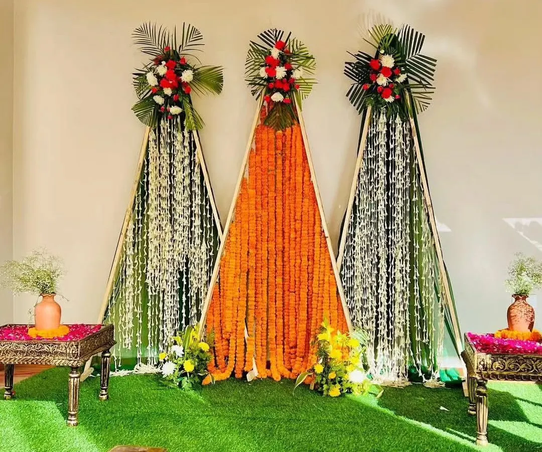 This is a modern and minimalist Haldi backdrop we created. The design uses three triangular frames with lush green turf as a base, featuring vibrant orange marigold curtains and white floral strings, topped with fresh flower bouquets for a clean and contemporary look.