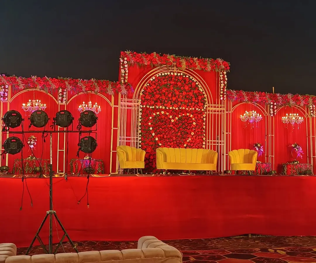 For a bold and royal wedding theme, I design grand stages like this one. It features a rich red floral backdrop with a heart motif, bright yellow velvet sofas for the couple, and dramatic lighting to create a memorable focal point.