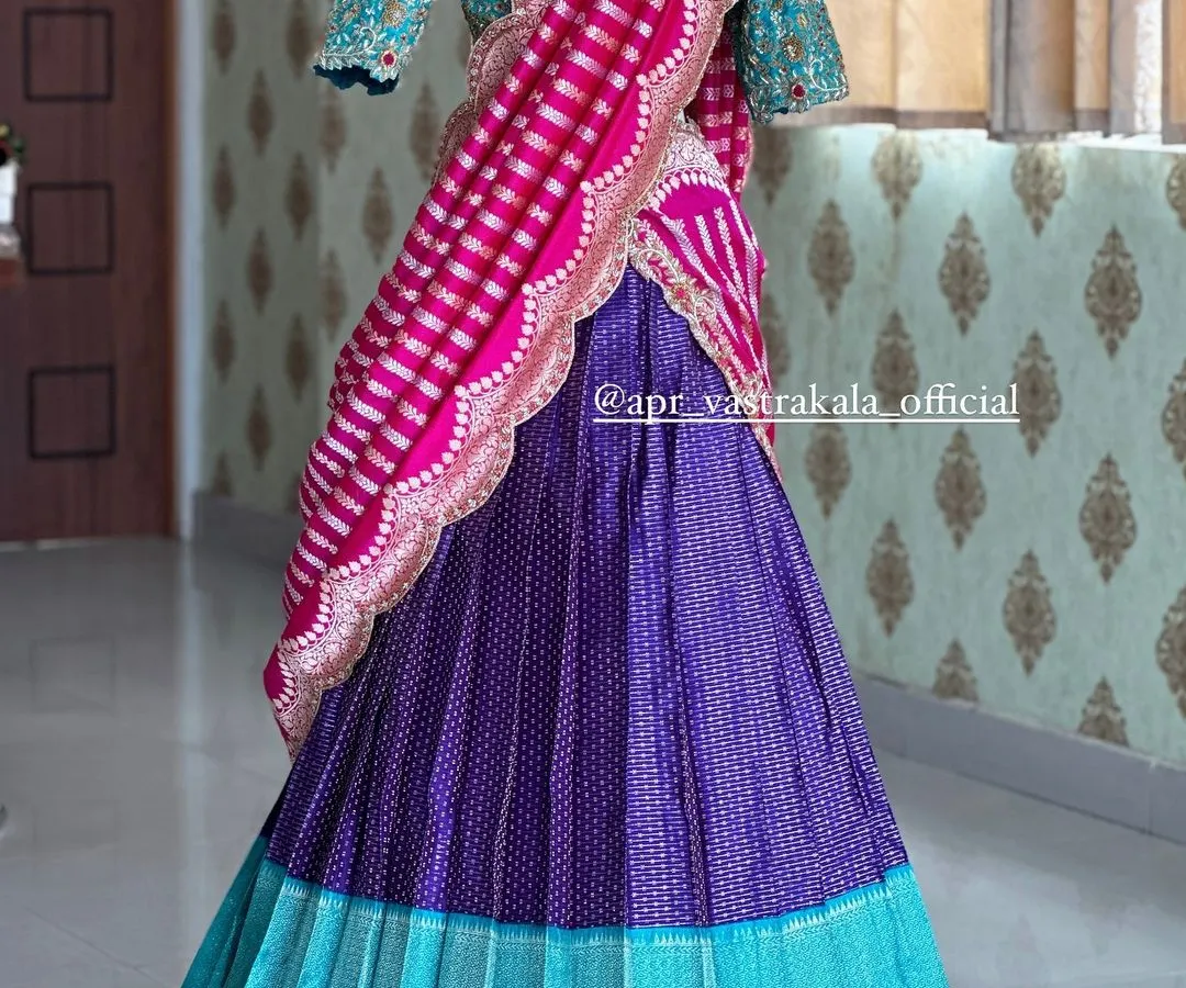 This bridal half-saree set is a masterpiece of color and texture. The design combines a pleated turquoise and purple brocade skirt with a heavily embroidered turquoise blouse and a contrasting striped pink dupatta for a bold, modern bridal look.