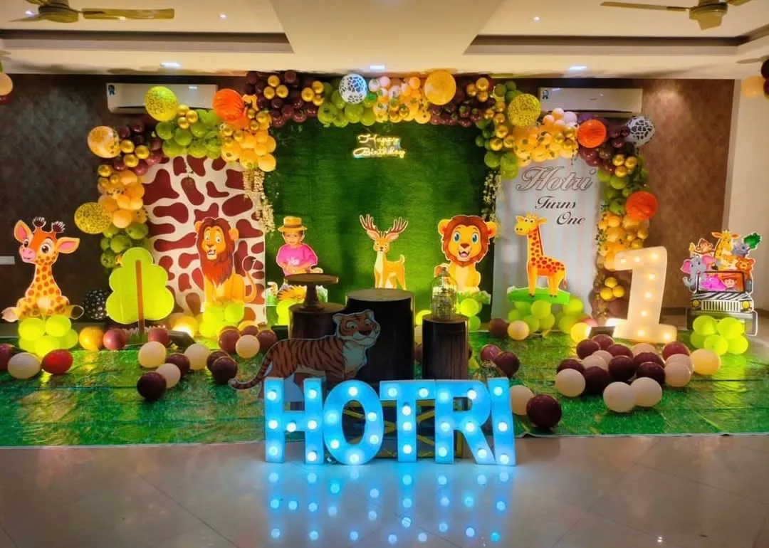 An elaborate jungle safari theme for a first birthday party. This large-scale setup includes a hedge wall backdrop, a custom giraffe-print panel, multiple animal cutouts, marquee letters for the name, and a '1' light-up prop, creating an immersive experience.
