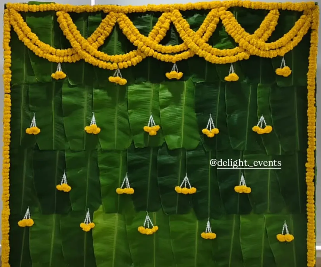 For an authentic and natural touch, we design beautiful backdrops like this one made entirely of fresh banana leaves. The simple marigold garlands and jasmine tassels make it perfect for home functions like a baby shower or pooja.