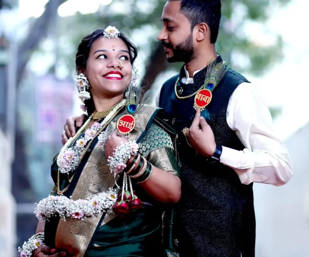 A happy couple shares a candid moment during their outdoor baby shower photoshoot. The mom-to-be is adorned in traditional floral jewelry, and they both wear playful 'Aai' (Mom) and 'Baba' (Dad) badges.