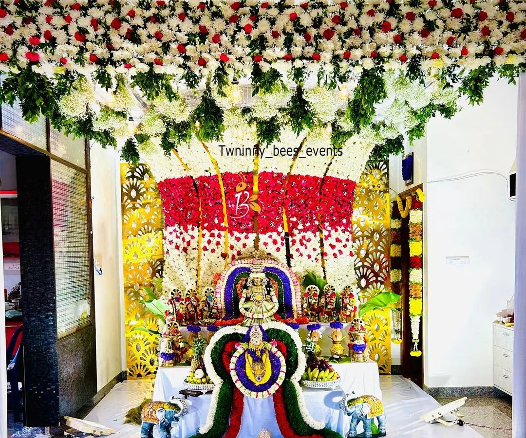 An elaborate floral mantap designed for a Varamahalakshmi puja at home. This decoration features a dense ceiling of white and red flowers flowing down into a multi-colored floral backdrop for the deity, creating a truly divine atmosphere.