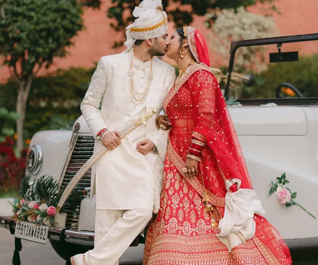 A moment of pure joy as the couple shares a kiss against a classic vintage car. The vibrant red of the bride's lehenga and the groom's elegant white sherwani create a cinematic portrait of their Punjabi wedding celebration.
