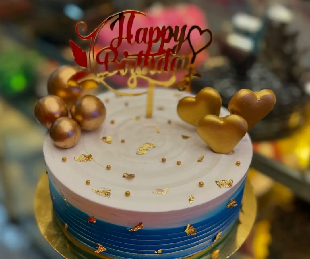 A birthday cake designed to make someone feel truly special. This creation features a beautiful blue and white gradient, adorned with golden hearts, spheres, and delicate gold leaf flakes, all topped with an elegant 'Happy Birthday' sign. I can create a similar masterpiece for your next celebration.