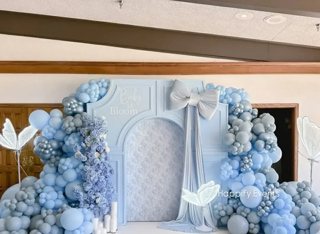 This premium "Baby in Bloom" baby shower backdrop features a sophisticated pastel blue theme. The decor includes a custom arched panel, a large fabric bow, and dense organic balloon garlands in varying shades of blue, accented with glowing butterfly props.