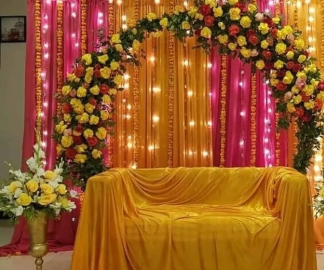 For pre-wedding functions like Haldi or Mehendi, I create vibrant backdrops that are perfect for photos. This setup features a beautiful arch of fresh yellow and red roses against rich pink and orange drapes, with fairy lights to add a warm glow.