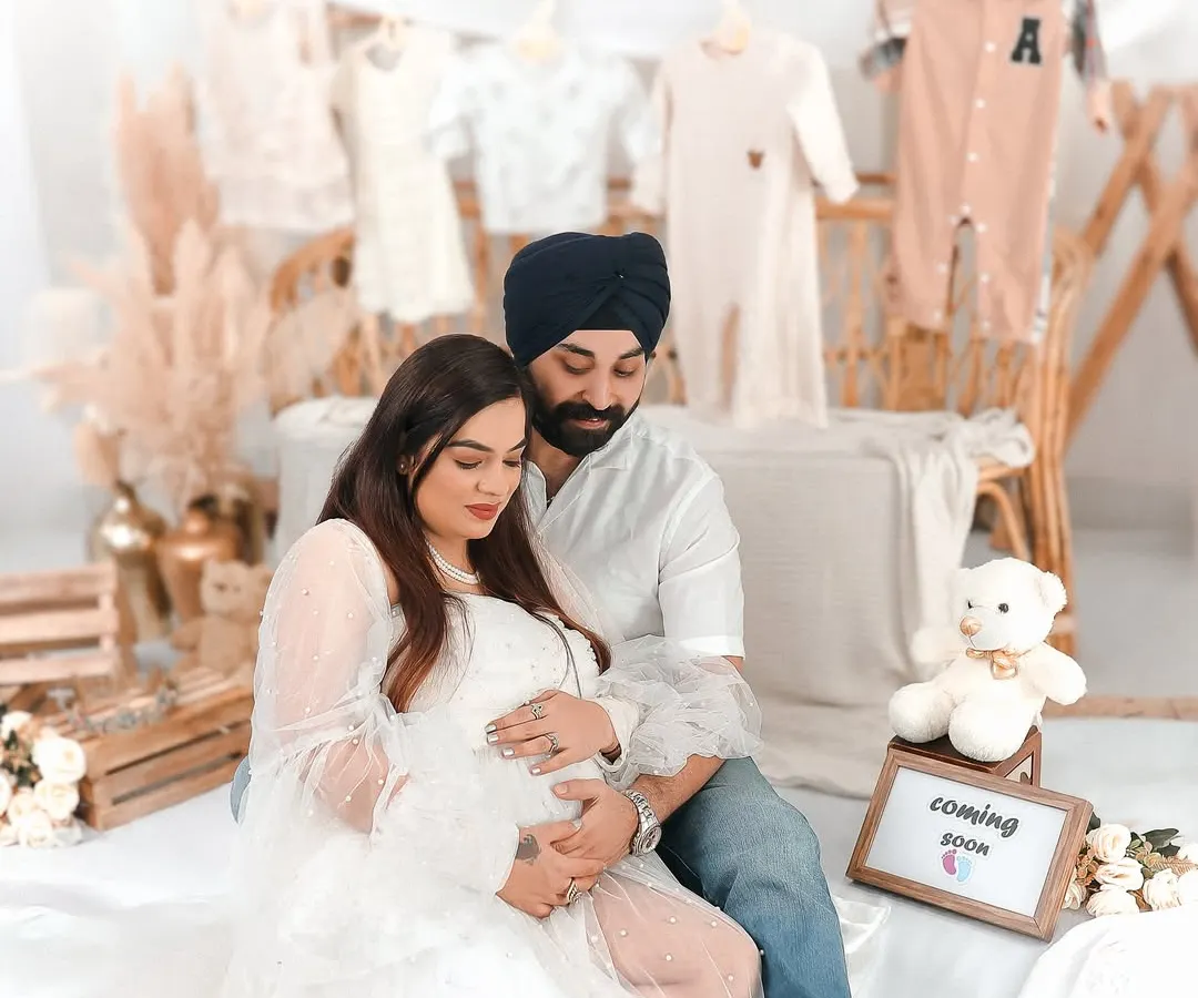 A tender moment between expectant parents during a studio maternity session. The soft, neutral backdrop with hanging baby clothes and a 'Coming Soon' sign creates a feeling of gentle anticipation and love.