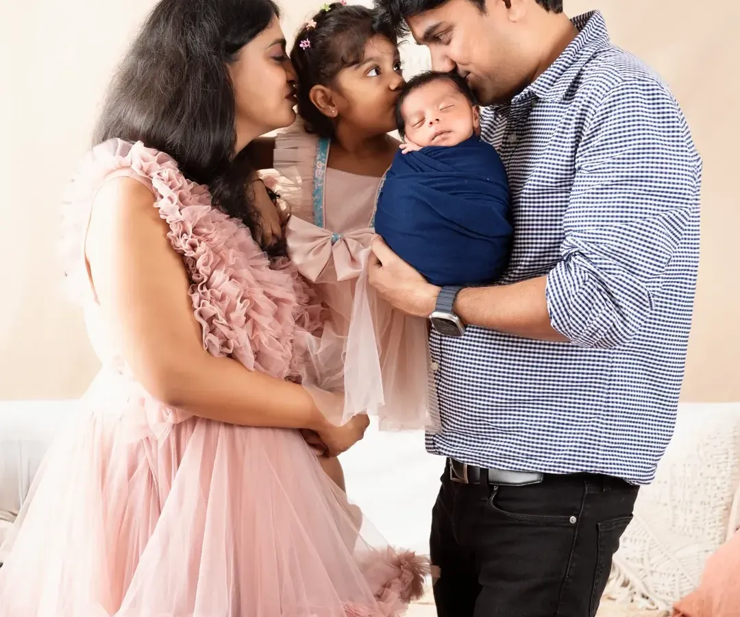 A heartwarming family portrait capturing a tender moment. The mother, father, and older sister share kisses with the newest family member, a newborn boy wrapped snugly in blue.