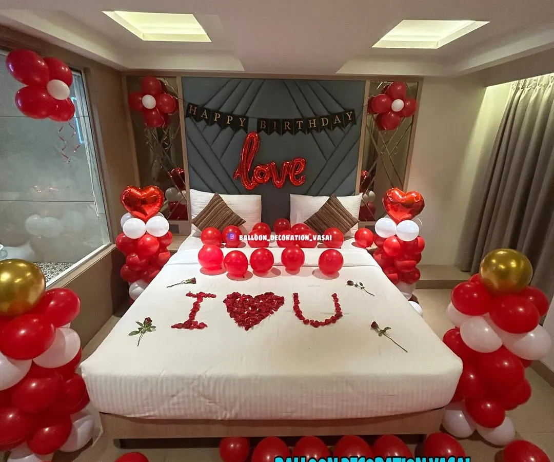 Surprise your partner with a classic romantic room decoration. This setup includes red and white balloon pillars, heart-shaped foil balloons, and a bed covered in rose petals spelling out "I ❤️ U" for a truly memorable birthday or anniversary celebration.