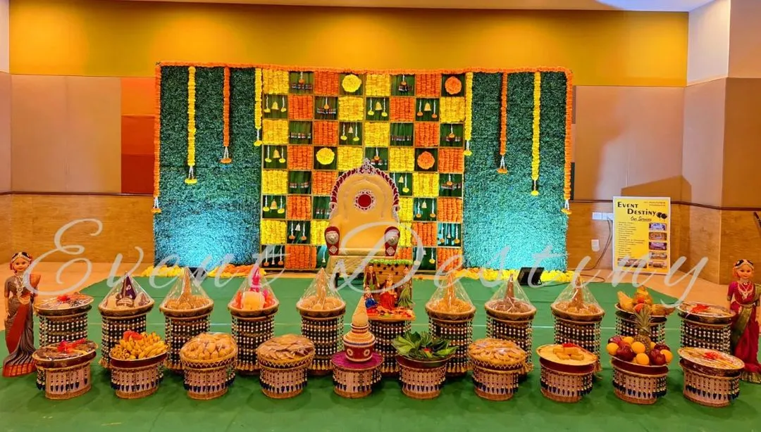 This is a complete setup for a half-saree function, showing both the main stage and the traditional seer plate arrangement. The backdrop features a unique checkerboard pattern of fresh marigolds and green leaves.