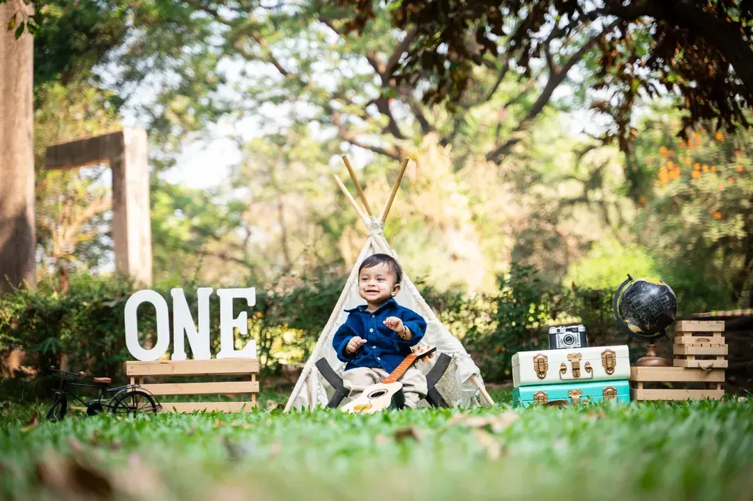 A beautifully styled outdoor first birthday photoshoot featuring a baby boy sitting in front of a teepee, surrounded by props like a 'ONE' sign, vintage suitcases, and a globe.