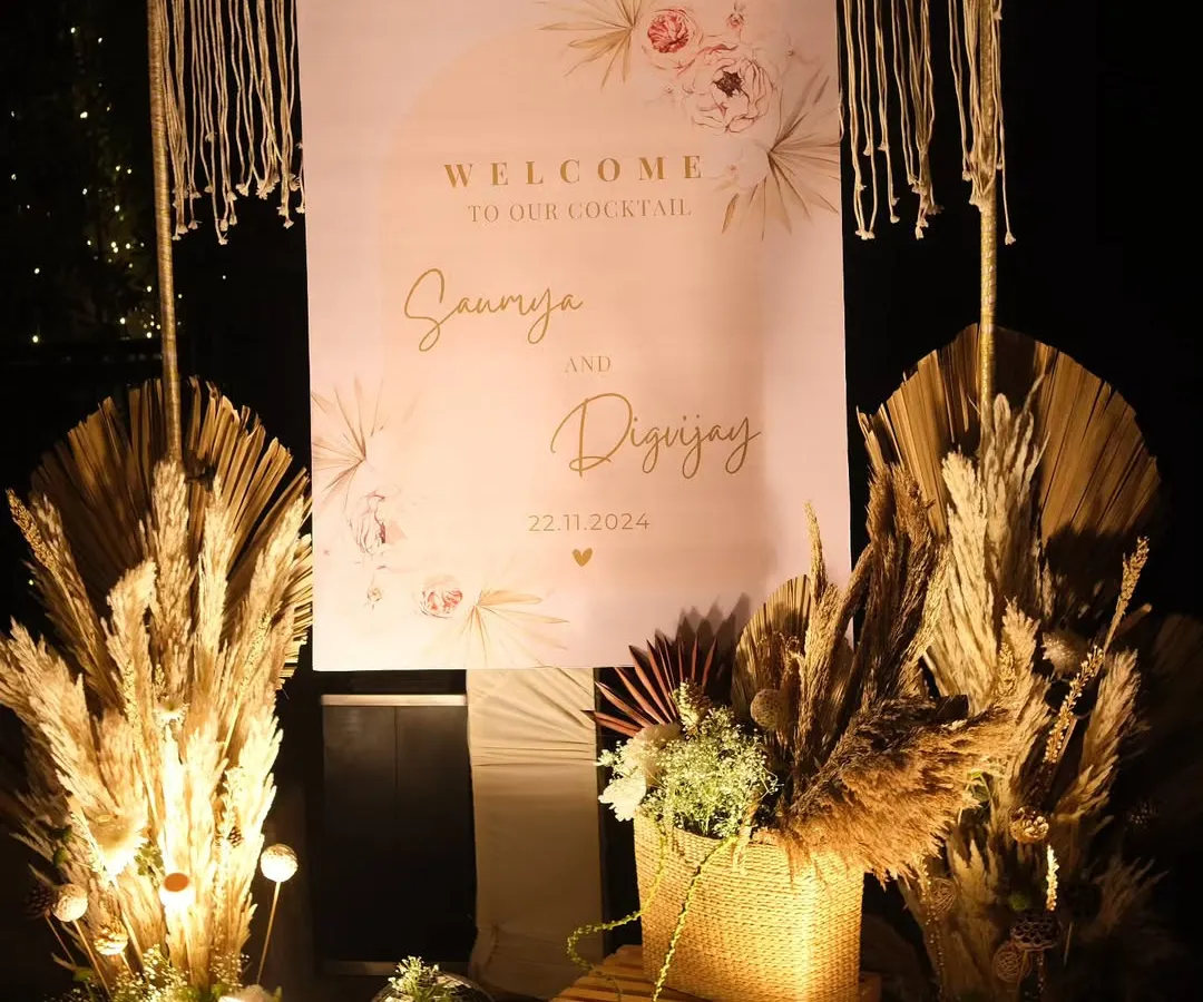 A personalized welcome sign for a boho-chic cocktail party. This setup combines elegant calligraphy with natural elements like pampas grass, dried palms, and a macrame frame to set a warm and stylish tone right from the entrance.