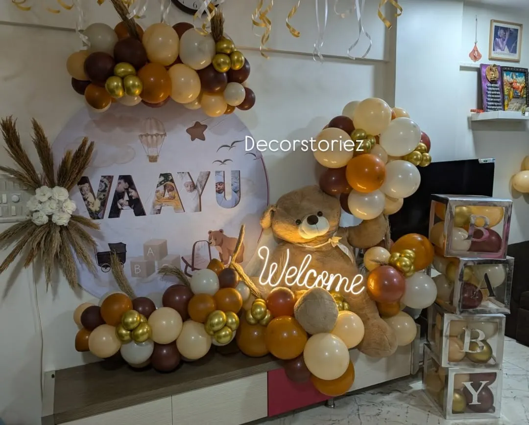 This modern, themed decoration was created to welcome baby Vaayu. We used an earthy palette of brown, beige, and gold balloons, a custom name backdrop, a large teddy bear, and a neon 'Welcome' sign for a stylish and personalized celebration.