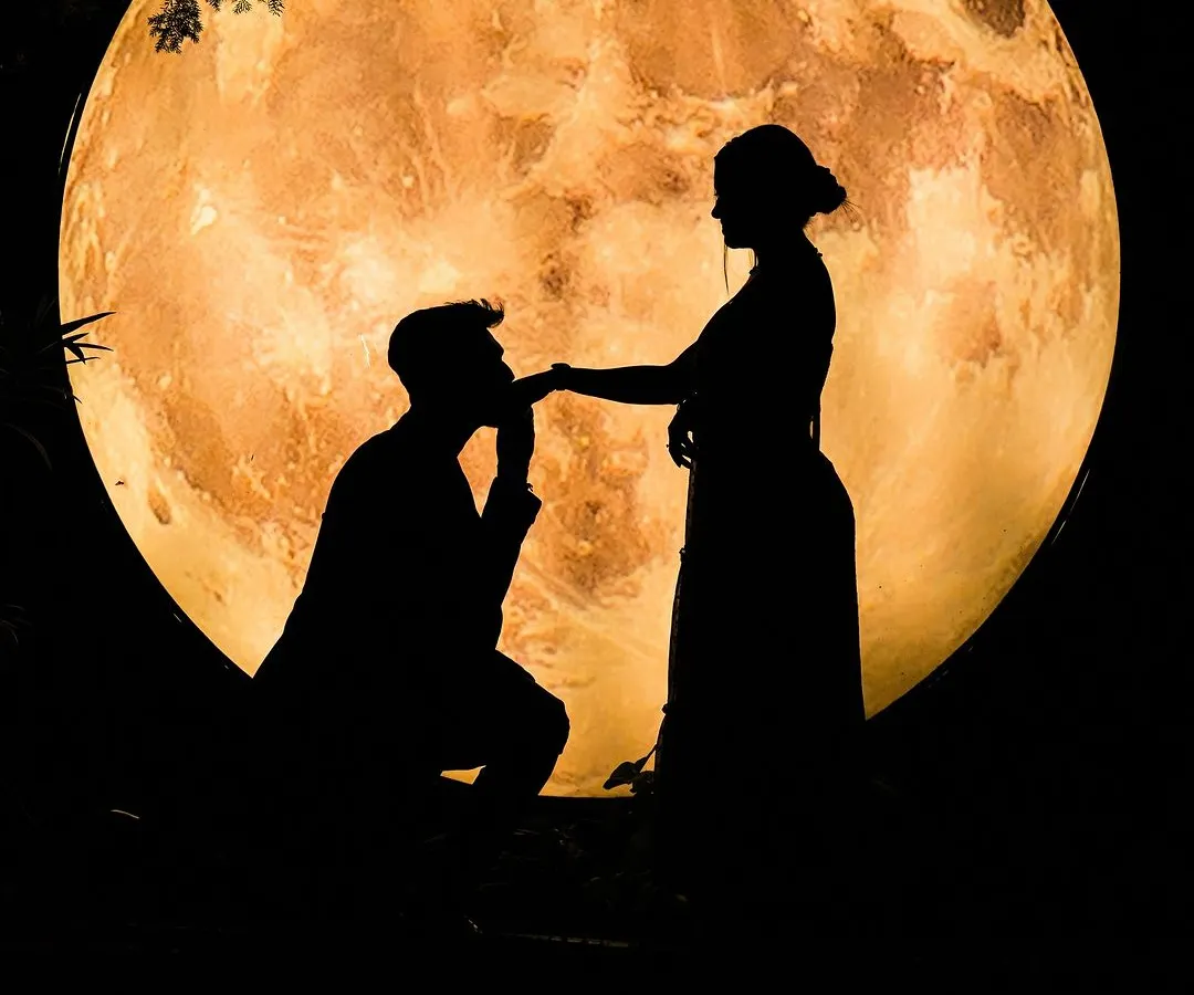 A classic romantic silhouette of a couple against a large, glowing moon prop, perfect for a save-the-date announcement or a timeless piece of art.