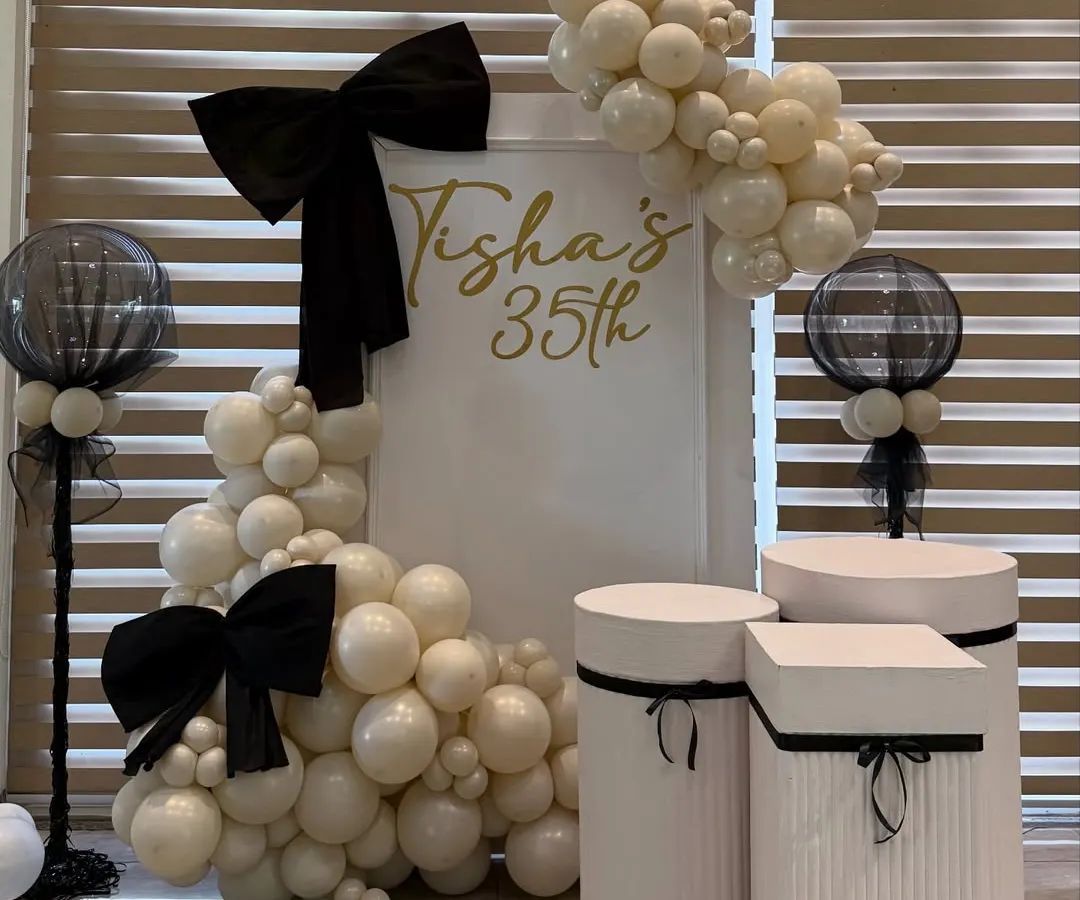 For a chic 35th birthday, we designed a sophisticated black, white, and gold theme. This setup features custom signage, elegant plinths for the cake display, and organic balloon garlands accented with statement bows for a touch of modern glamour.