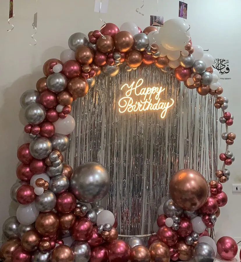 This is how we bring the party to you. We created this luxe balloon arch for a special birthday celebration at home, using a mix of metallic rose gold, silver, and white balloons, complete with a 'Happy Birthday' neon sign against a silver fringe curtain.