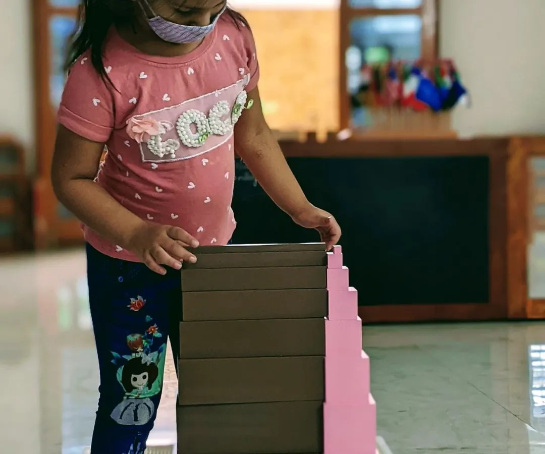 Deep concentration is a hallmark of Montessori learning. This child is fully engaged with the Pink Tower and Broad Stair, classic sensorial materials that help develop visual discrimination of size and dimension.