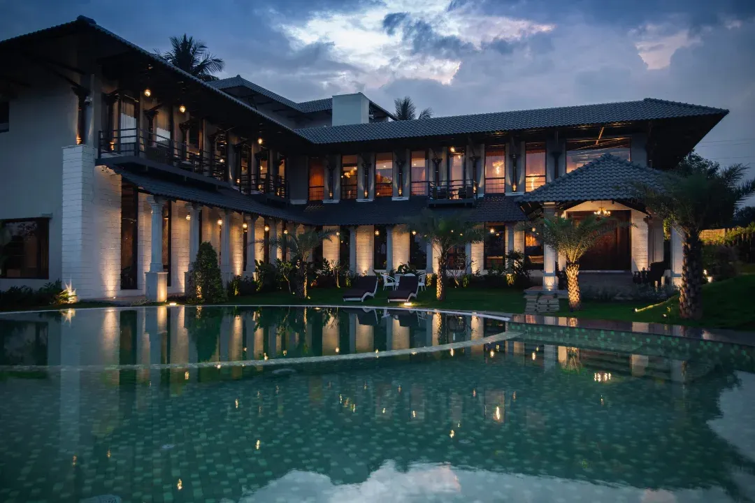 We design homes that exist in harmony with their surroundings. This farmhouse exterior showcases a seamless blend of modern architecture and tranquil nature, with a serene pool that mirrors the sky and the structure's elegant form, creating a private resort-like escape.
