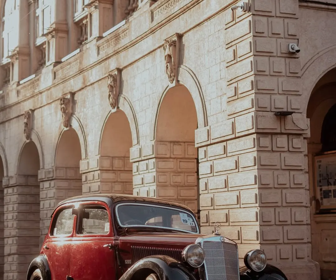 A classic two-tone burgundy and black Mercedes-Benz sedan from a bygone era, parked on a city street. The warm evening light highlights its vintage design against the grand, stone architecture of a heritage building in Mumbai.