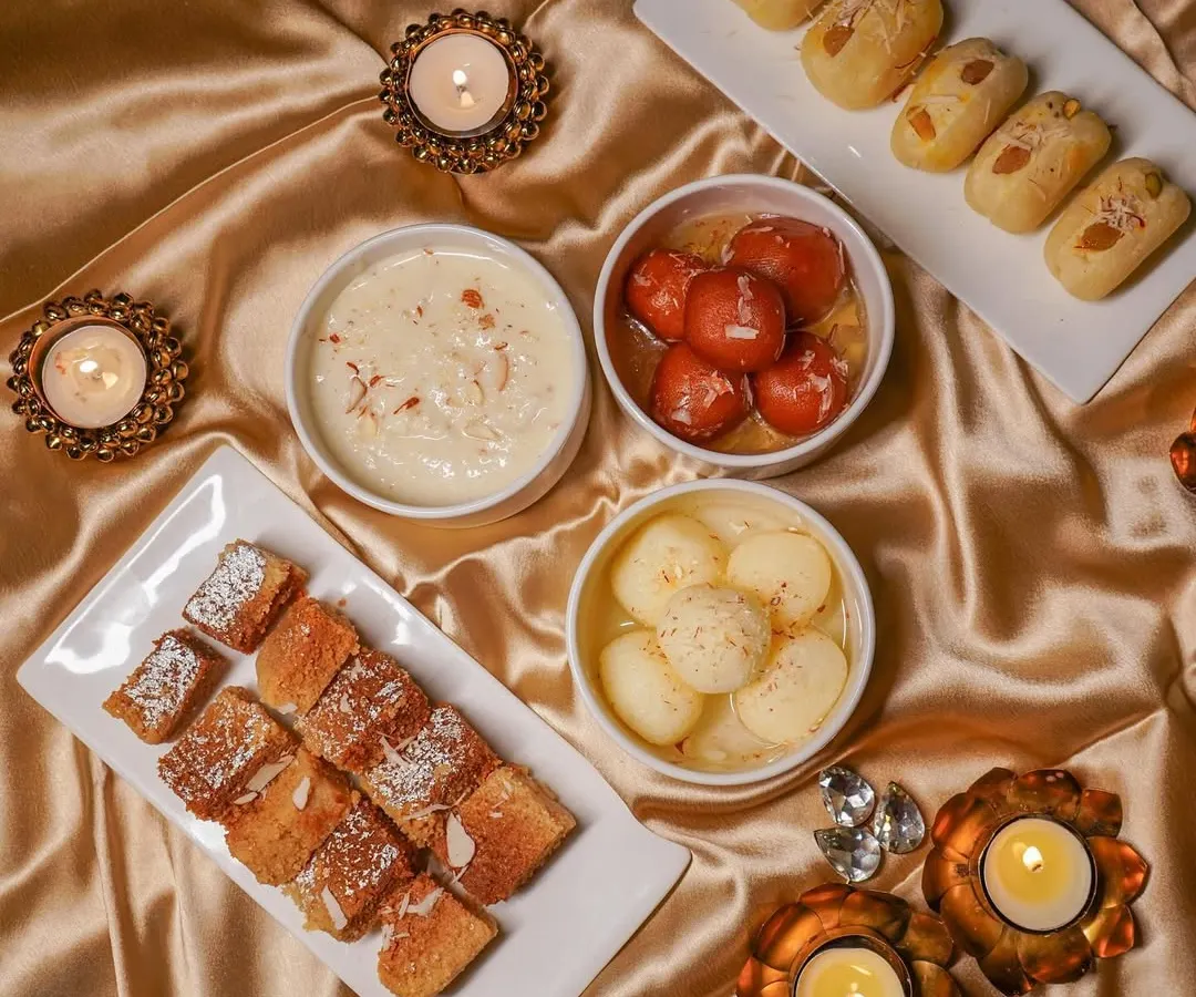 A festive tabletop arrangement of Indian sweets, including gulab jamun and rasgulla, styled with golden fabric and lit diyas. This is an example of my food photography for seasonal campaigns and brand promotions.